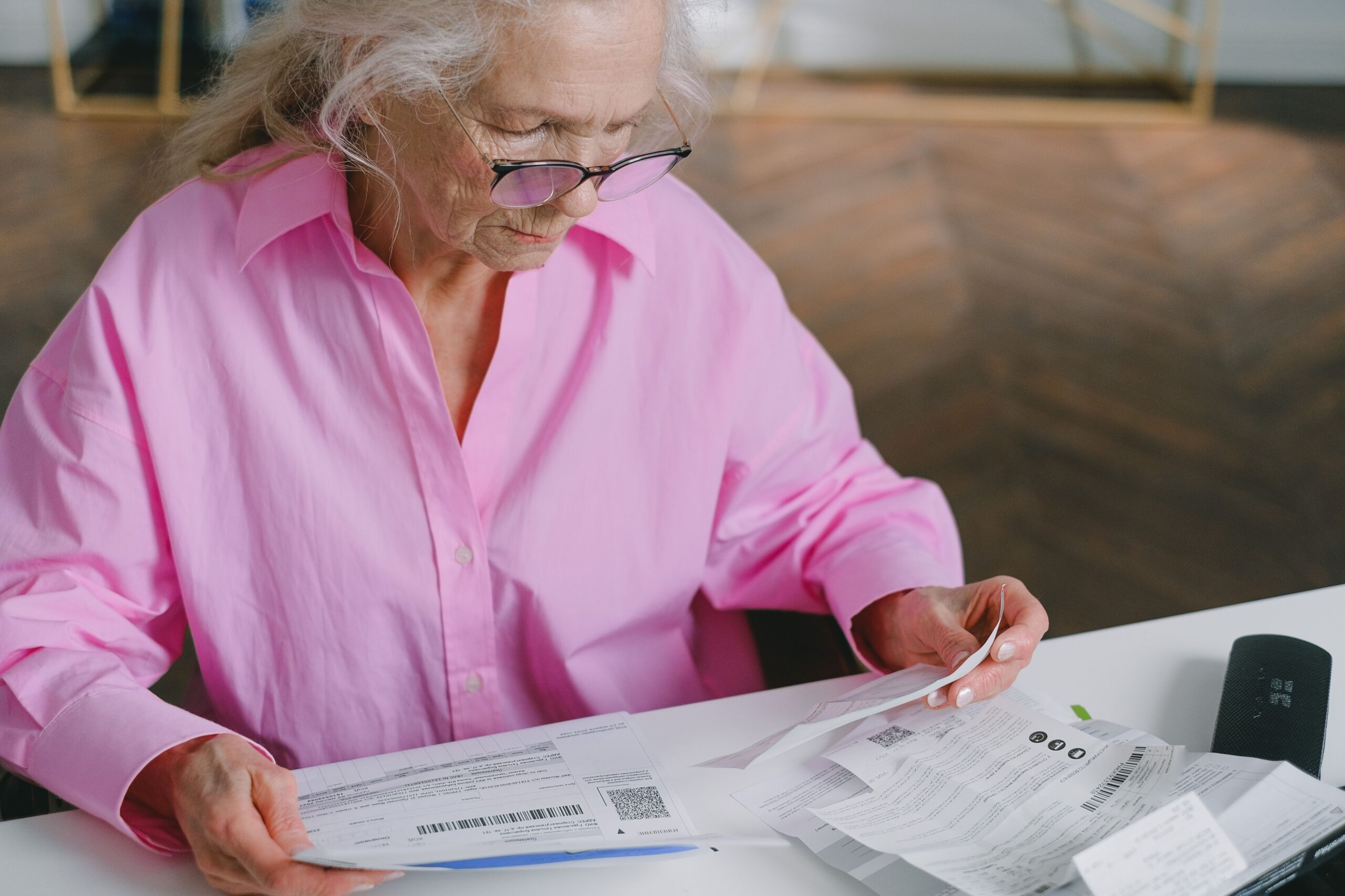 Home Elderly woman in pink blouse reading documents at a table indoors.